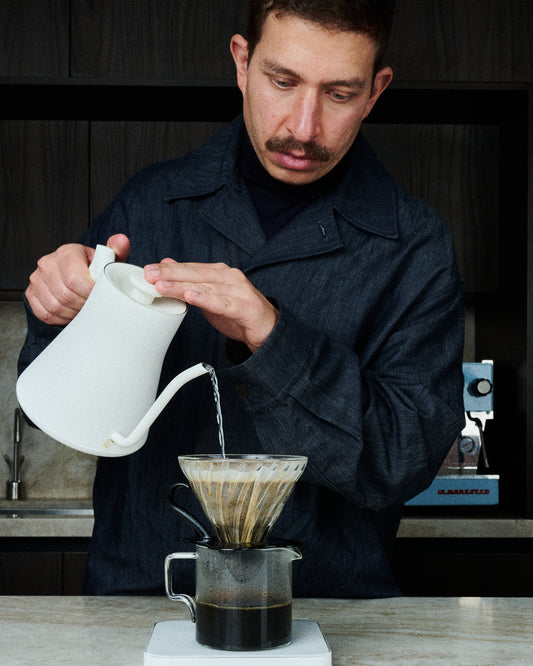 A man pouring hot water from a white gooseneck kettle into a pour-over coffee dripper set over a glass server